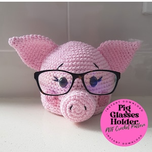 May include: A pink crocheted pig wearing glasses. The pig is a glasses holder. The image includes the text "Instant Download Pig Glasses Holder PDF Crochet Pattern Instant Download".