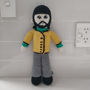 May include: A crocheted doll with black hair, a yellow jacket with black buttons, green sleeves, gray pants, and black shoes.