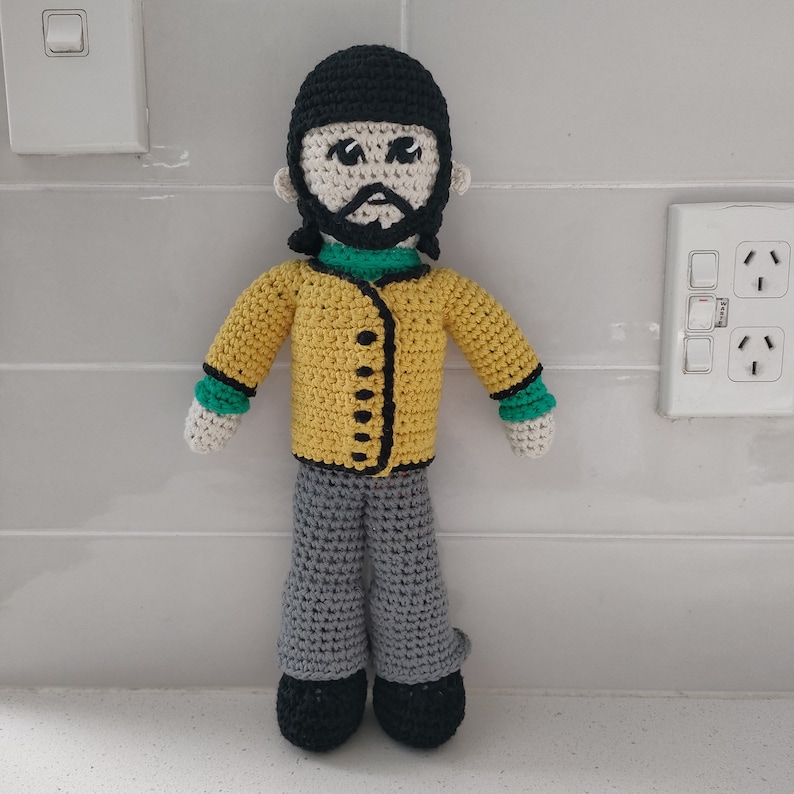 May include: A crocheted doll with black hair, a yellow jacket with black buttons, green sleeves, gray pants, and black shoes.
