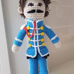 May include: Crocheted doll with brown hair, a white face, a black mustache, and a blue and white uniform with yellow buttons. The doll is wearing brown shoes.