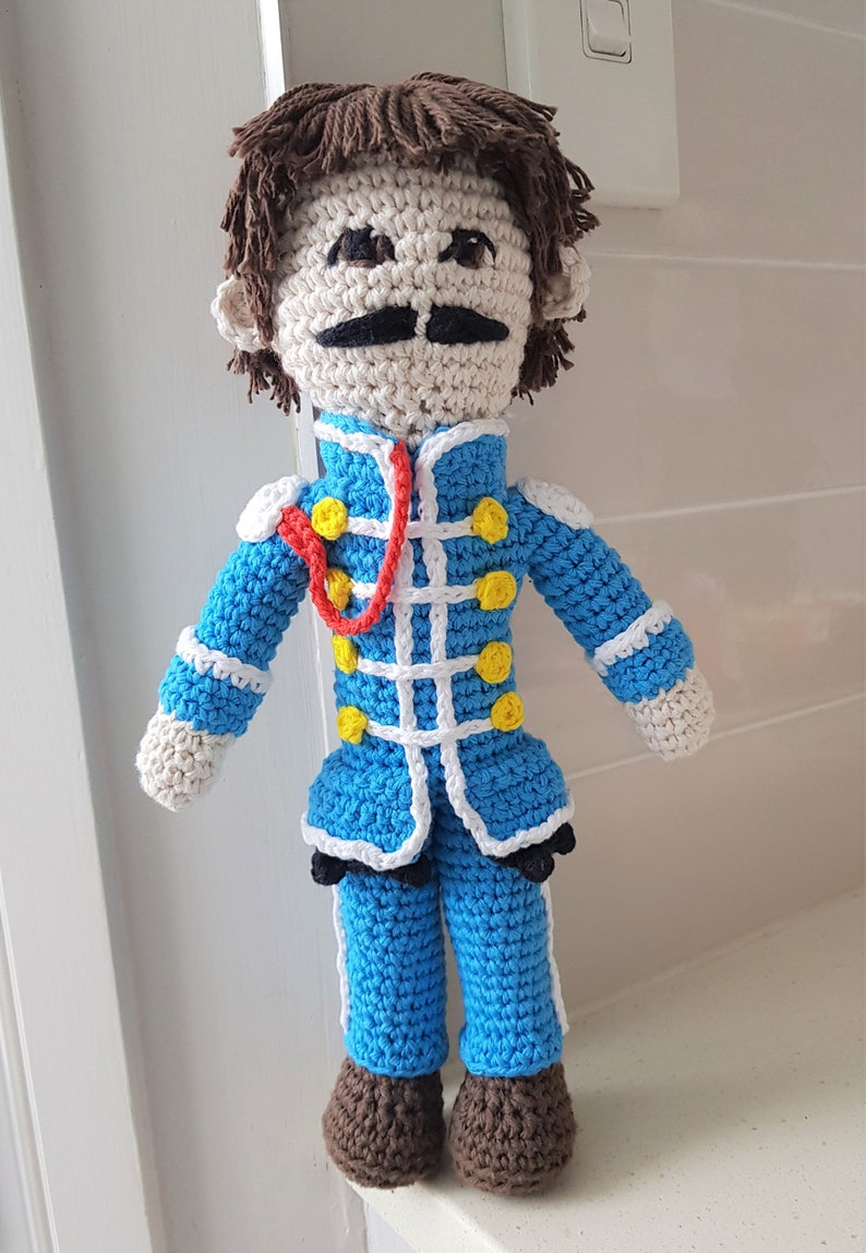 May include: Crocheted doll with brown hair, a white face, a black mustache, and a blue and white uniform with yellow buttons. The doll is wearing brown shoes.