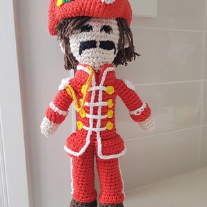 May include: A crocheted doll wearing a red and white uniform with a yellow sash and a red hat with a green leaf. The doll has brown hair, a white face, and a black mustache.