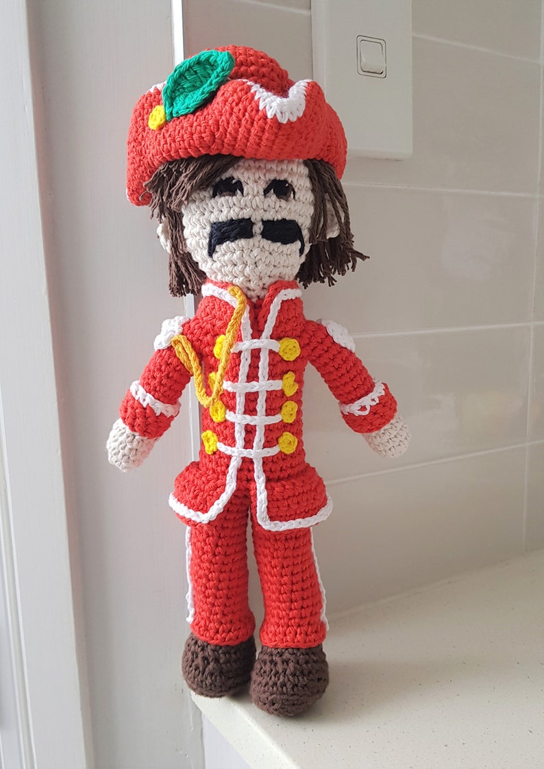 May include: A crocheted doll wearing a red and white uniform with a yellow sash and a red hat with a green leaf. The doll has brown hair, a white face, and a black mustache.