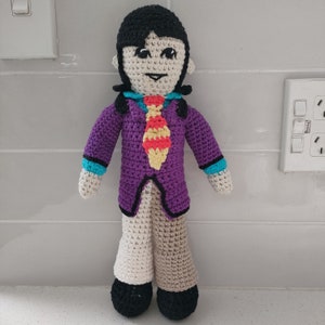 May include: Crocheted doll with black hair, purple jacket, yellow and red tie, and beige pants. The doll is standing on a white tiled surface.