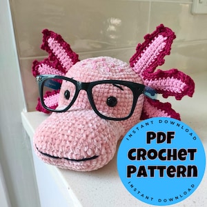 May include: A handmade pink crochet axolotl wearing black-framed glasses. The axolotl has dark pink gills and a smiling mouth. A blue circle with the text "PDF CROCHET PATTERN" is in the lower right corner.