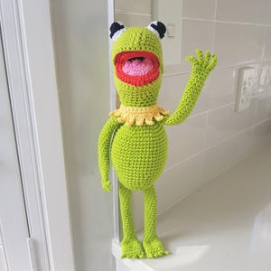 May include: A handmade, green crochet frog doll with a wide open mouth, red lips, and a pink tongue. The frog has black and white eyes, a yellow collar, and is posed with one arm raised. The doll is standing on a white surface.