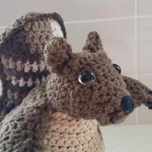 Puede incluir: Un juguete de ardilla hecho a mano con ganchillo, en tonos marrones, beige y blanco. La ardilla tiene ojos grandes y claros y una nariz y patas azul oscuro. La cola está rayada con marrón y blanco. El juguete está hecho con un punto de ganchillo texturizado.