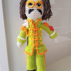 May include: Crocheted doll with brown hair, yellow glasses, a black mustache, and a green, orange, and white outfit.