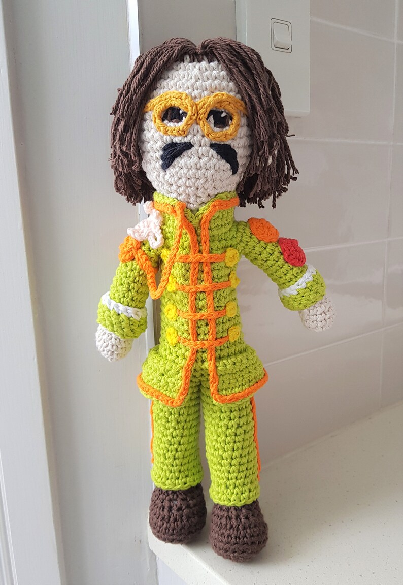 May include: Crocheted doll with brown hair, yellow glasses, a black mustache, and a green, orange, and white outfit.