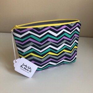 May include: A rectangular cosmetic bag with a colorful chevron pattern in black, white, gray, teal, yellow, and purple. The bag has a yellow zipper and a tag that reads "Looks Like She Made It HANDCRAFTED GOODS."