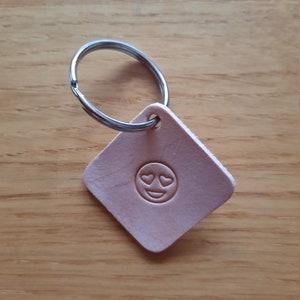 May include: A light brown leather keychain with a silver key ring. The keychain has a square shape and features a heart-eyed emoji embossed on the front.