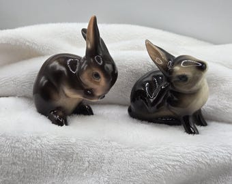 May include: Two ceramic rabbit figurines. The rabbits are brown and tan with glossy finishes. One rabbit is upright with its ears perked up, and the other is in a seated position, grooming itself. The figurines are on a white, textured surface.