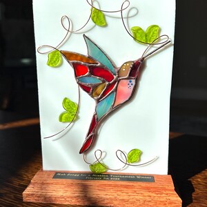 May include: A stained glass hummingbird sculpture with a colorful design of red, blue, orange, and pink. The hummingbird is mounted on a white background with copper wire vines and green glass leaves. The base is a wooden plaque with text.