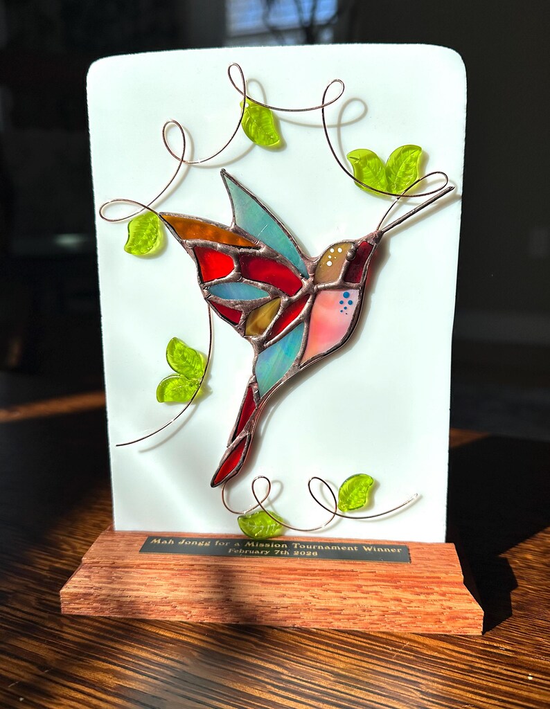 May include: A stained glass hummingbird sculpture with a colorful design of red, blue, orange, and pink. The hummingbird is mounted on a white background with copper wire vines and green glass leaves. The base is a wooden plaque with text.