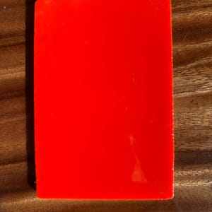 May include: A solid red rectangular object with a glossy finish. The object is set against a wooden surface with visible grain. The lighting creates shadows, highlighting the object's shape and texture.