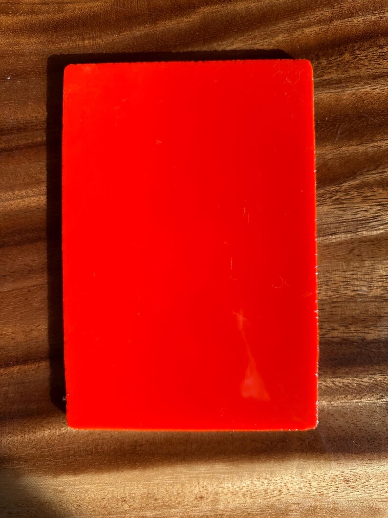 May include: A solid red rectangular object with a glossy finish. The object is set against a wooden surface with visible grain. The lighting creates shadows, highlighting the object's shape and texture.