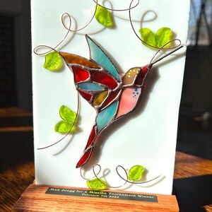 May include: A stained glass hummingbird sculpture with a white background and wooden base. The bird features red, blue, yellow, and pink glass. Copper wire swirls and green glass leaves surround the bird. The base has text that reads "Mah Jongg for a Mission Tournament Winner February 7th 2020."