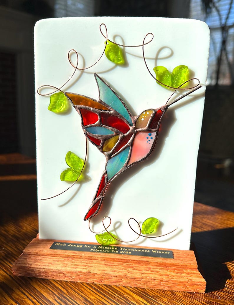May include: A stained glass hummingbird sculpture with a white background and wooden base. The bird features red, blue, yellow, and pink glass. Copper wire swirls and green glass leaves surround the bird. The base has text that reads "Mah Jongg for a Mission Tournament Winner February 7th 2020."