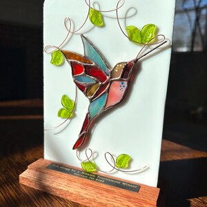May include: A stained glass hummingbird with red, blue, and orange wings is mounted on a white rectangular plaque. Copper wire vines and green glass leaves surround the bird. The plaque sits on a wooden base with an engraved plate.