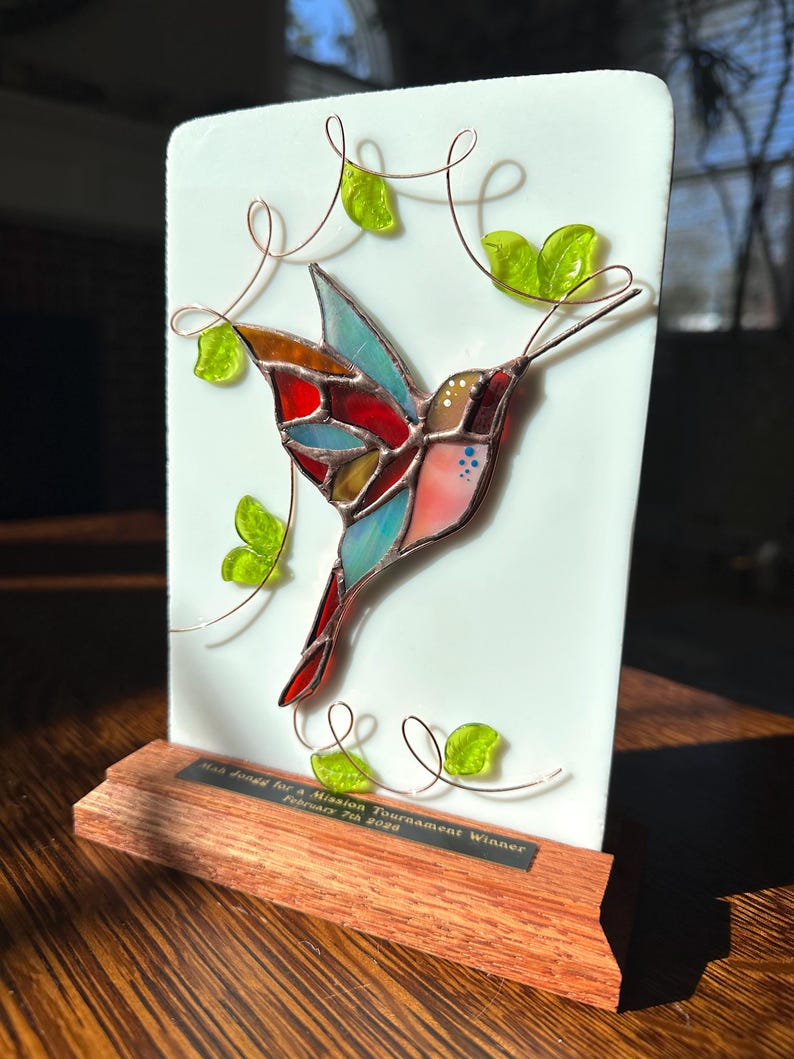 May include: A stained glass hummingbird with red, blue, and orange wings is mounted on a white rectangular plaque. Copper wire vines and green glass leaves surround the bird. The plaque sits on a wooden base with an engraved plate.