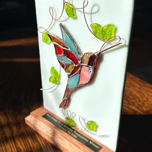 May include: A stained glass hummingbird sculpture with a colorful design, mounted on a wooden base. The hummingbird has red, blue, and yellow accents. Green glass leaves and copper wire vines decorate the white background. The base has text that reads "Mission Tournament Winner".