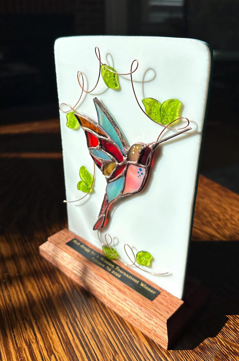 May include: A stained glass hummingbird sculpture with a colorful design, mounted on a wooden base. The hummingbird has red, blue, and yellow accents. Green glass leaves and copper wire vines decorate the white background. The base has text that reads "Mission Tournament Winner".
