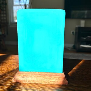 May include: A rectangular, bright turquoise acrylic panel is mounted on a stained, wooden base. The panel has rounded corners and stands upright. The wooden base is a warm brown color. The item is displayed on a wooden table.