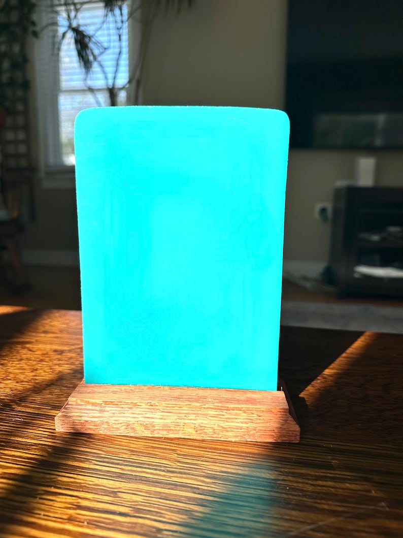 May include: A rectangular, bright turquoise acrylic panel is mounted on a stained, wooden base. The panel has rounded corners and stands upright. The wooden base is a warm brown color. The item is displayed on a wooden table.