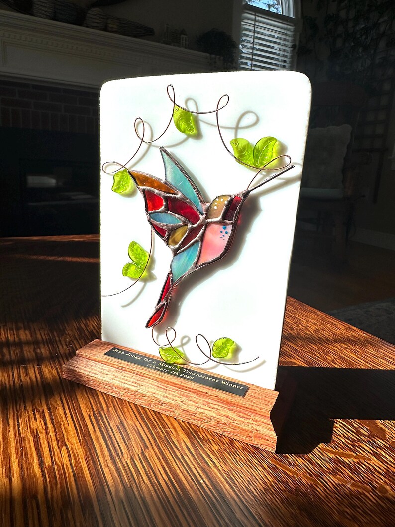 May include: A stained glass hummingbird with red, blue, and pink wings, mounted on a white background. The hummingbird is set on a wooden base. The text on the base reads: "Mah Jongg for a Mission Tournament Winner February 7th 2020."
