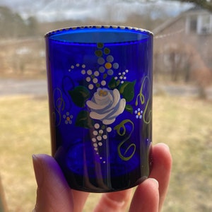 May include: A cobalt blue glass shot glass with a white rim and hand-painted floral design. The design features a white rose with green leaves and white dots.