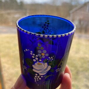 May include: A cobalt blue glass shot glass with a white floral design. The glass has a white rim and a white dot pattern around the top.