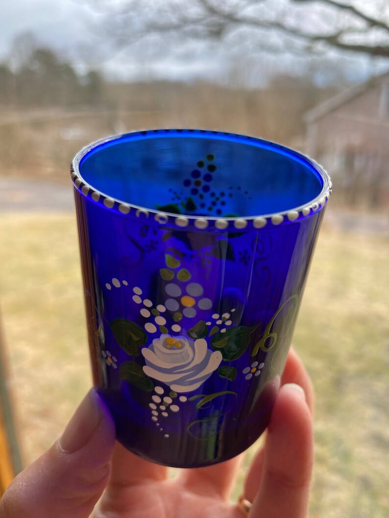 May include: A cobalt blue glass shot glass with a white floral design. The glass has a white rim and a white dot pattern around the top.