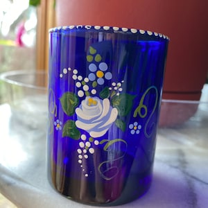 May include: A cobalt blue glass tumbler with a white rim and hand-painted floral design. The design features a white rose with blue and yellow accents, green leaves, and white dots.