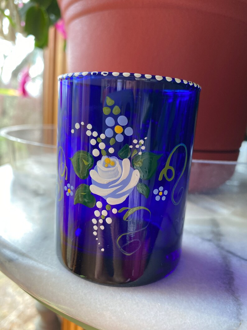 May include: A cobalt blue glass tumbler with a white rim and hand-painted floral design. The design features a white rose with blue and yellow accents, green leaves, and white dots.