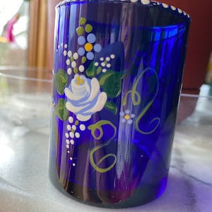 May include: A cobalt blue glass tumbler with a white rim and hand-painted floral design. The design features a white rose with blue and yellow accents, green leaves, and white dots.