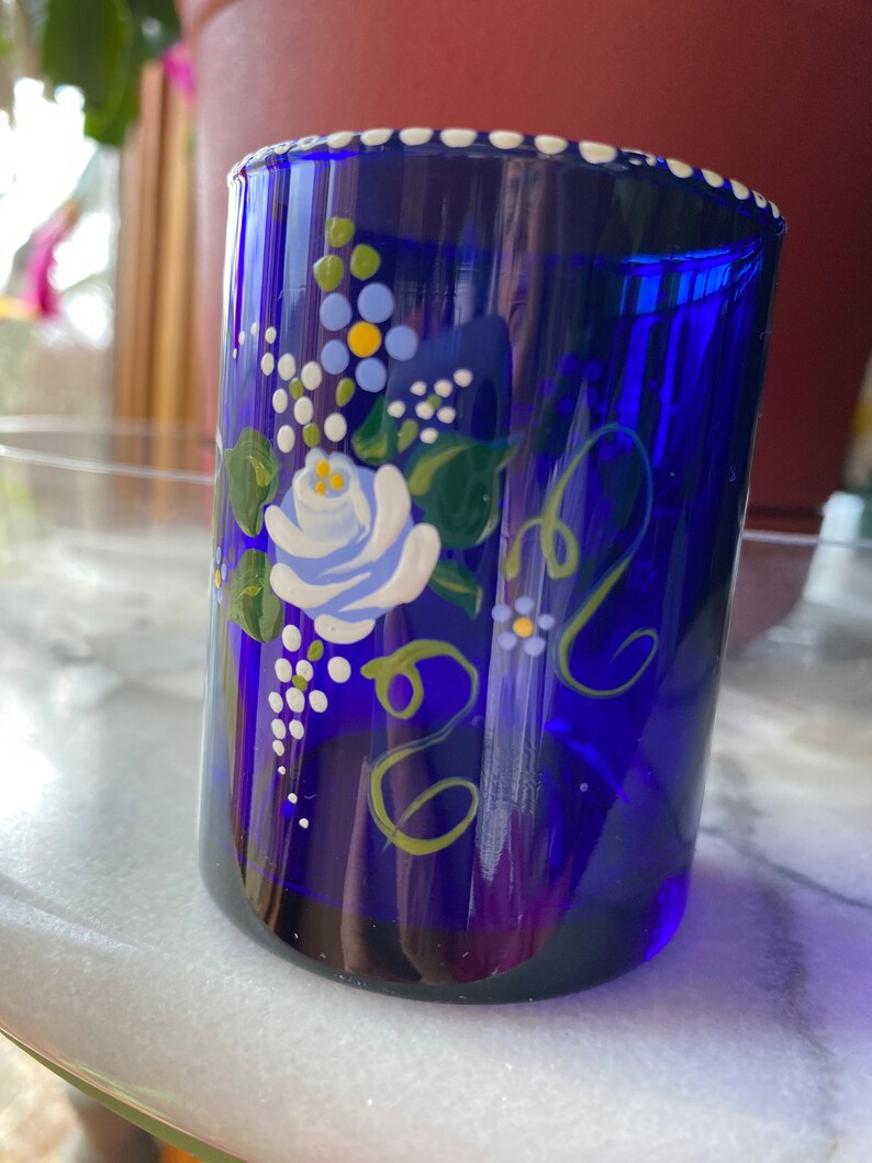 May include: A cobalt blue glass tumbler with a white rim and hand-painted floral design. The design features a white rose with blue and yellow accents, green leaves, and white dots.