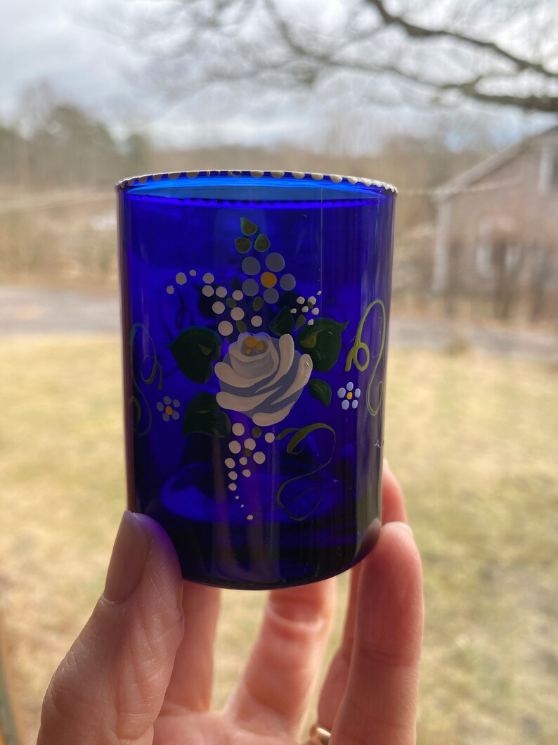 May include: A cobalt blue glass shot glass with a white, blue, and green floral design painted on the side.