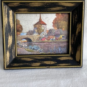 May include: A framed painting of a quaint village scene with a clock tower, bridge, and houses. The painting is done in a watercolor style with muted colors and a soft, dreamy feel.