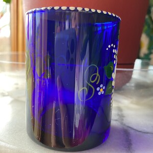 May include: A cobalt blue glass cup with a white rim and white polka dots. The cup is hand-painted with green leaves and white flowers.