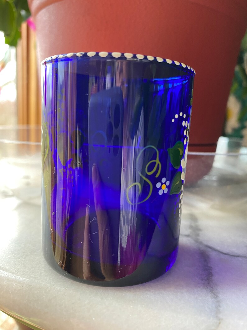 May include: A cobalt blue glass cup with a white rim and white polka dots. The cup is hand-painted with green leaves and white flowers.