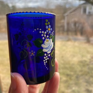 May include: Cobalt blue glass shot glass with a white rose and floral design painted on the side.