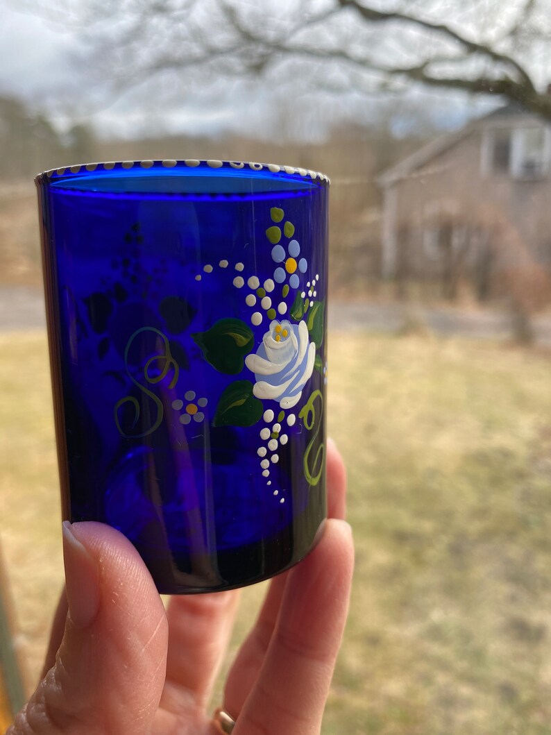 May include: Cobalt blue glass shot glass with a white rose and floral design painted on the side.