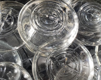 May include: A close-up of a stack of clear glass jar lids with a raised, textured design. The lids are stacked on top of each other, creating a pattern of overlapping circles.