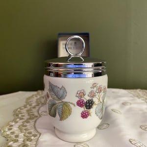 May include: A white porcelain egg coddler featuring a botanical pattern of green leaves, white flowers, and clusters of red and black blackberries. The container has a polished silver-tone metal lid with a circular ring handle on top.