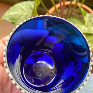 May include: A small, cobalt blue glass cup with a silver rim and a beaded edge. The cup has a white floral design on the inside.
