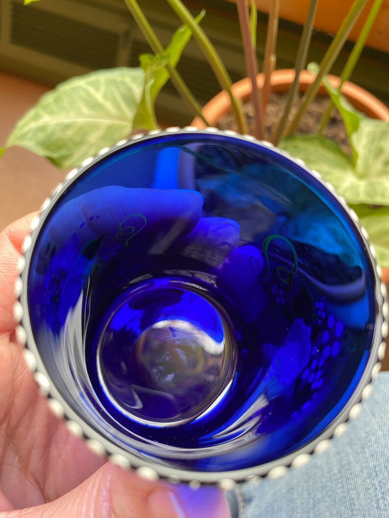 May include: A small, cobalt blue glass cup with a silver rim and a beaded edge. The cup has a white floral design on the inside.