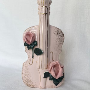 May include: A pink ceramic violin-shaped vase with gold accents. The vase features two pink roses with green leaves. The violin's strings and edges are highlighted with gold detailing. A decorative piece for home decor.