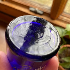 May include: Cobalt blue glass shot glass with white and gold floral design. The glass has a clear, reflective surface.