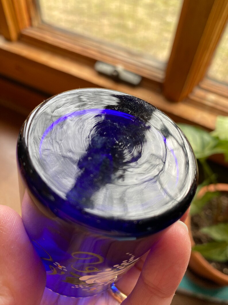 May include: Cobalt blue glass shot glass with white and gold floral design. The glass has a clear, reflective surface.