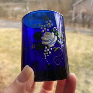May include: A cobalt blue glass shot glass with a hand-painted floral design. The design features a white rose with green leaves and white dots.